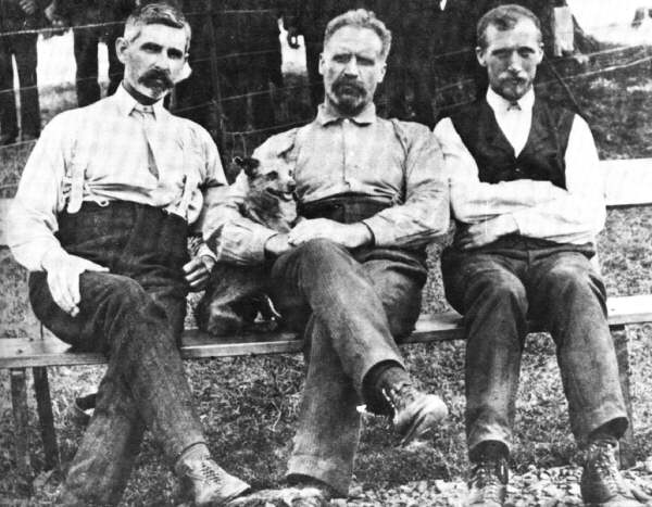 an old photograph showing 3 bearded men seated on a bench with the man in the center with his arm around a small dog